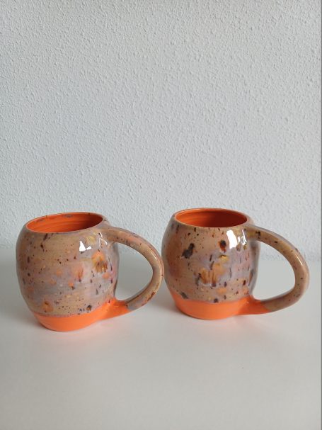 Twee aardewerk mokken met een oranje onderkant en speckled patroon.